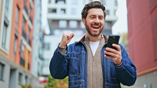 Individual radiates happiness and triumph while engaging with mobile phone. Moment of success captured amidst modern city buildings, showing pure joy.