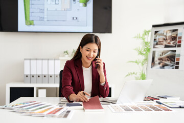 Concentrated young interior designer woman sitting at table with blueprint drawing ,swatches and material , discussing interior design with client by phone, choosing materials for decorations.