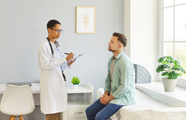 Fototapeta premium Portrait of female doctor talking with young male patient sitting on couch in clinic in exam room. Physician giving consultation to a man during medical examination in office. Medicine concept.