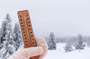 Frosty winter weather, snowfall, low temperature concept. Thermometer in hand wearing warm knitted wool mitten against background of snow-covered natural landscape with fir trees in snow