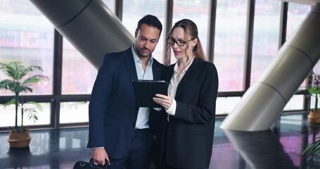 Business professionals gathered around tablet in glass hallway of office building, discussing and sharing ideas, upper body frontal view - Powered by Adobe