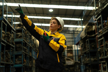 Hispanic female foreman worker holding walkie talkie, checking inventory of auto parts and engine maintenance duties in factory.