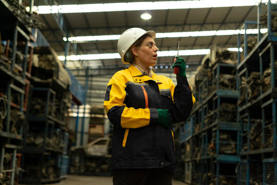 Hispanic female foreman worker holding walkie talkie, checking inventory of auto parts and engine maintenance duties in factory.