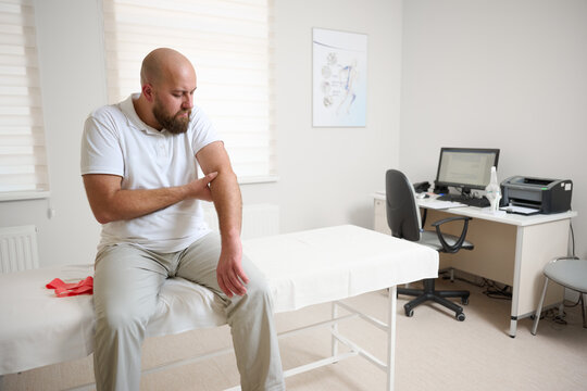Man suffering from elbow pain during medical consultation in physiotherapy clinic. Patient holding arm in discomfort while doctor examines his shoulder. Concept of joint injury,