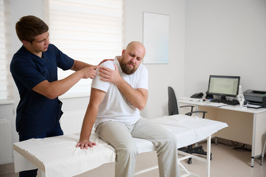 Physiotherapist examining patient shoulder joint during rehabilitation therapy session in modern medical clinic. Man receiving orthopedic treatment and physical therapy for injury recovery.