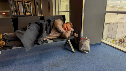 A weary traveler, Caucasian male, dozes on an airport bench, embodying the spontaneous serenity of Lost Luggage Day