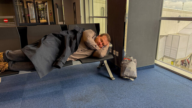 A weary traveler, Caucasian male, dozes on an airport bench, embodying the spontaneous serenity of Lost Luggage Day - Powered by Adobe