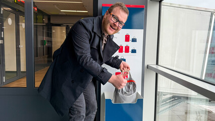 A jovial Caucasian man in a sleek coat retrieves parcels from a bustling, modern post-machine as if unwrapping gifts on Cyber Monday or Singles' Day, infusing mundane moments with seasonal cheer