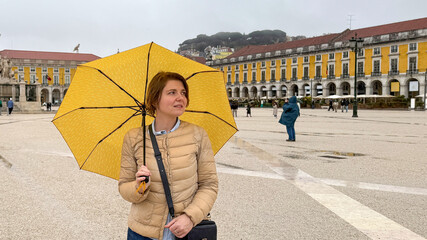 A woman with auburn hair and fair skin stands under a yellow umbrella in a historical plaza,...