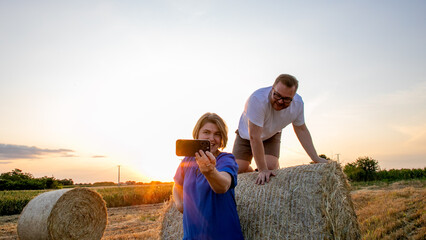 In golden twilight, a Caucasian man and woman capture bucolic joy atop sunlit hay bales, echoing harvest festival camaraderie, celebrating Lughnasadh