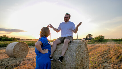 In a sunlit rural tableau, a Caucasian man and woman share laughter atop hay bales, celebrating Lughnasadh and summer's whimsical end