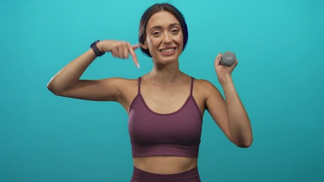 Young hispanic woman pointing to toned arm while holding a 1kg dumbbell in a teal studio; confidence wellness health.
