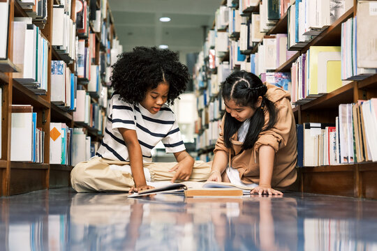 Two diverse children sit on the floor between bookshelves in a library, reading and sharing a book. A joyful academic moment filled with kids literature, knowledge, and classroom curiosity.