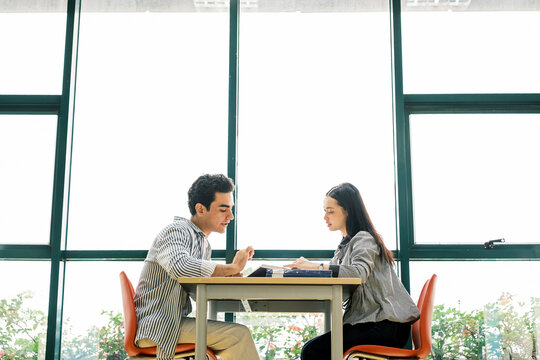 Two students sit across a table by large windows, collaborating on paperwork in a bright modern room. Scene captures teamwork, partnership, study, coaching, seminar prep, and campus meeting vibes. - Powered by Adobe