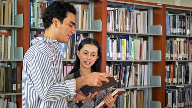 Caucasian man and Asian woman students smile while browsing and discussing books in a library aisle, holding open volumes among shelves, showing learning, reading, and academic collaboration.