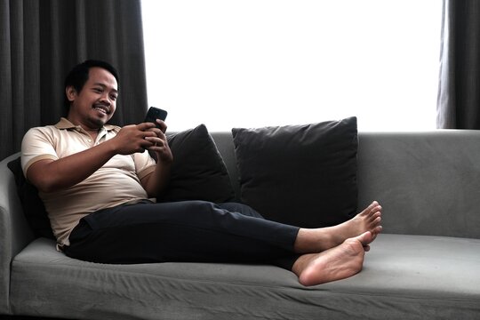 Southeast Asian man in a beige shirt lying comfortably on a sofa, smiling while using his smartphone in a cozy indoor setting.