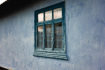 Old blue wooden window with cracked paint and lace curtains on a textured wall in a rustic setting. moldova