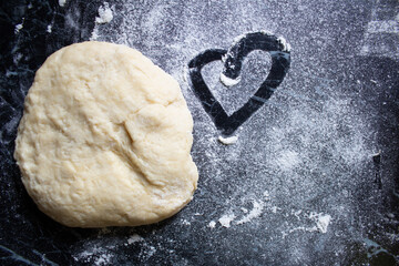 Raw dough on dark table with scattered flour and a heart drawn in it. Romantic rustic baking scene. Top view, homemade cooking concept, cozy kitchen atmosphere.