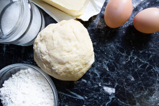 Raw dough and classic baking ingredients — flour, eggs, butter, sugar — on a dark rustic table. Top view. Homemade cooking and pastry preparation concept.