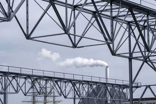 Industrial scene showing a tall smokestack releasing steam and vapor into the sky, framed by metal infrastructure and structural elements under dense cloud cover in a modern industry setting