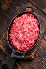 Raw ground wild game meat (venison/boar) in a black skillet on a rustic wooden board, seasoned with fresh thyme and peppercorns.