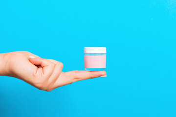 Female hand holding cream bottle of lotion isolated. Girl give jar cosmetic products on blue...