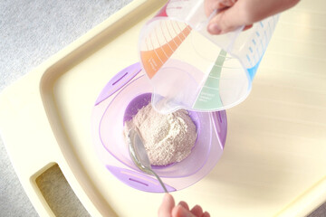 baby food and nutrition concept - mother hands with jar, spoon and bowl preparing cereal from powder close up