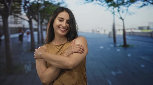Young hispanic woman hugging her bare shoulders with arms crossed on urban street, smiling gently; self care contentment.