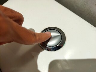 A finger presses the split chrome flush button on a white toilet tank, indicating activation of a dual-flush system.