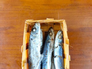 A woven basket holds several small, cooked or dried fish with silver and blue hues, resting on a warm wooden surface. Ikan pindang from a local market in Malang, Indonesia.