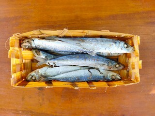 A woven basket holds several small, cooked or dried fish with silver and blue hues, resting on a warm wooden surface. Ikan pindang from a local market in Malang, Indonesia.