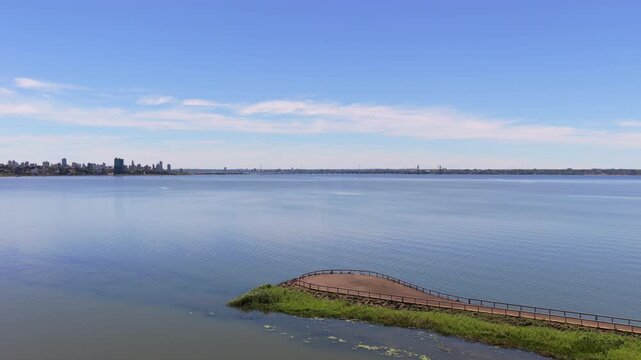 Aerial over Paran&aacute; River at Miguel Lan&uacute;s, Posadas: curved walkway on green bank, water expanse, and distant skyline under blue sky in Misiones, Argentina.