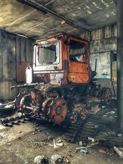 Fototapeta premium Abandoned Soviet-era hangar with a rusty tracked tractor, peeling paint, broken windows, scattered parts, and darkened concrete walls lit by side daylight.
