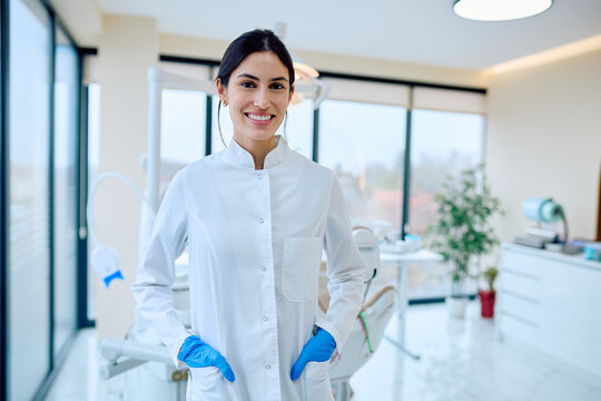 Young woman dentist wearing a white lab coat and blue gloves, smiling confidently in bright dental office - Powered by Adobe