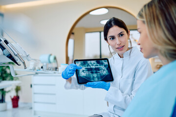 Dentist in white coat and gloves showing panoramic dental x-ray on digital tablet to a female patient during consultation