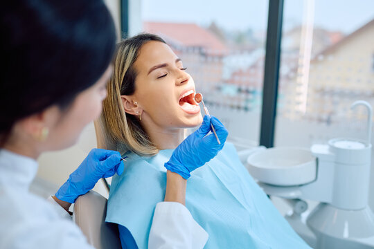 Female dentist wearing blue gloves examining a young woman's open mouth with dental tools during a routine checkup in a clinic