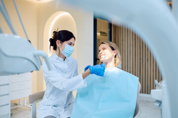 Dentist wearing protective mask and gloves holding teeth shade guide, comparing it with the client's healthy white smile in modern clinic