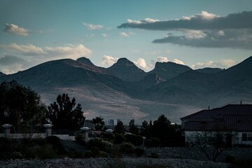 Scenic view of rugged mountain peaks under a moody sky with silhouetted houses and trees in the foreground at dusk