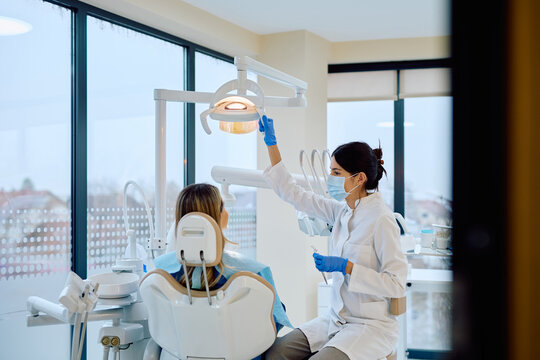 Female dentist wearing face mask and gloves adjusting examination lamp, preparing for a patient's routine dental examination in clinic - Powered by Adobe