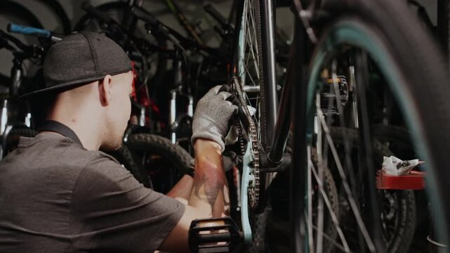 Young Caucasian technician wearing baseball cap repairing bicycle drivetrain system in workshop