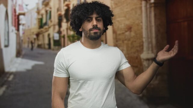 Man with curly hair shows open palm gesture and exposed forearm on a cobbled street in old town; subtle confidence.