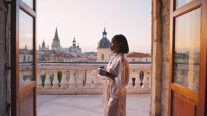 Obraz premium Young woman in silk robe standing on old european balcony with morning coffee at soft pastel sunrise light