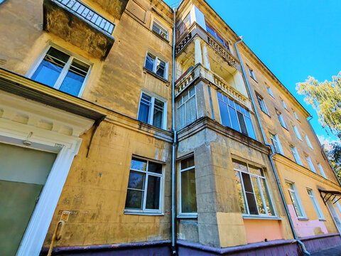 Old yellow residential building with worn balconies and decorative columns showing signs of plaster damage and weathering. The photo highlights facade aging and partial restoration, suitable for