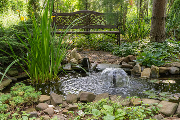 Serene garden pond with small waterfall, surrounded by rocks, lush greenery, and tall plants. Wooden bench is placed nearby, offering peaceful spot to relax amidst nature.