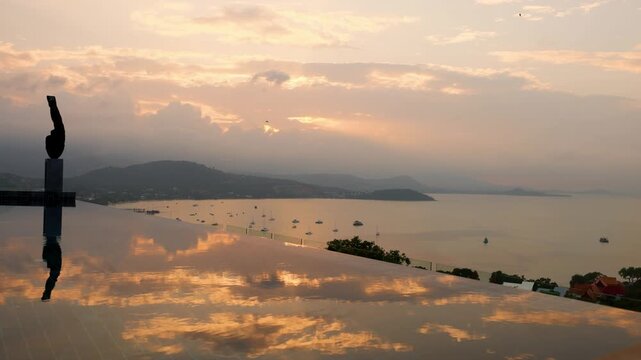 Tropical sunset timelapse over a calm bay with boats anchored near the shore, viewed from an infinity pool reflecting the golden sky. The serene scene captures luxury, tranquility, and tropical beauty