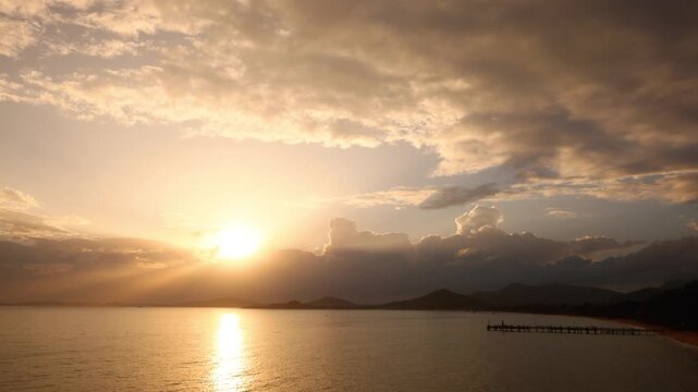 Golden tropical sunset over the sea with dramatic clouds, soft sunlight rays, and a silhouette of a pier extending into the calm water. The peaceful atmosphere and warm tones evoke relaxation
