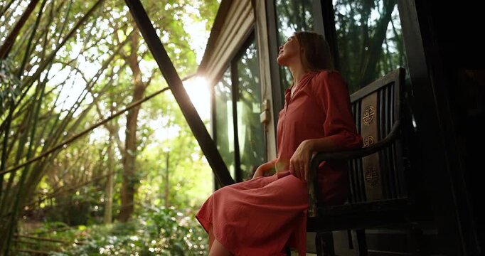 Woman in a coral dress relaxing on a wooden chair outside a cabin in the forest, enjoying sunlight streaming through bamboo trees. The scene conveys peace, mindfulness, and connection with nature in a