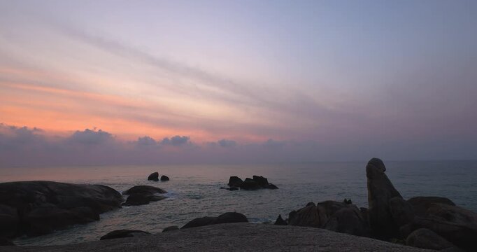 Tropical coastal timelapse of a serene sunset with soft pink and purple clouds over the calm sea and rocky shoreline. The tranquil scene captures the beauty of twilight and the peaceful rhythm of ocea