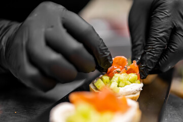 A chef in black gloves prepares gourmet appetizers, adding toppings to small toasts with cucumber and cream cheese spread.