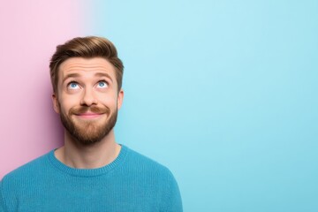 A man with a beard looks upwards, wearing a blue sweater, against a pink and blue background.